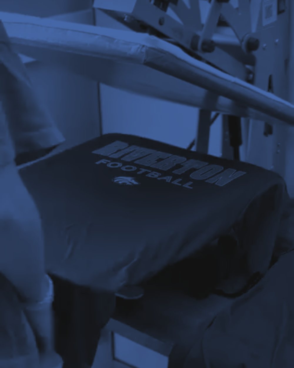 A black Riverton Football tee on a heat press in the Lift Threadz shop, with a shop worker's arm at the left edge of the frame.