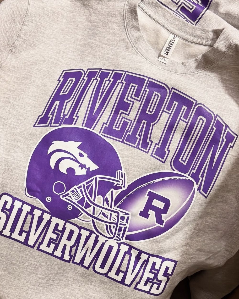 Heather oatmeal crewneck printed with a purple and white Riverton Silverwolves football helmet design, with matching crewnecks stacked behind it.